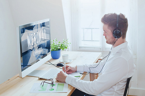 Man wearing headphones sitting at desk looking at a webinar on the screen