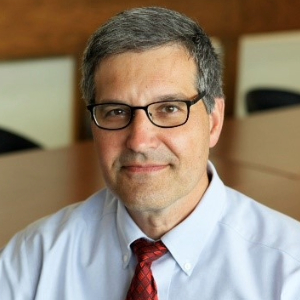 Short-haired person with relaxed, pleasant facial expression wearing a light button up shirt, red tie and rectangular glasses.