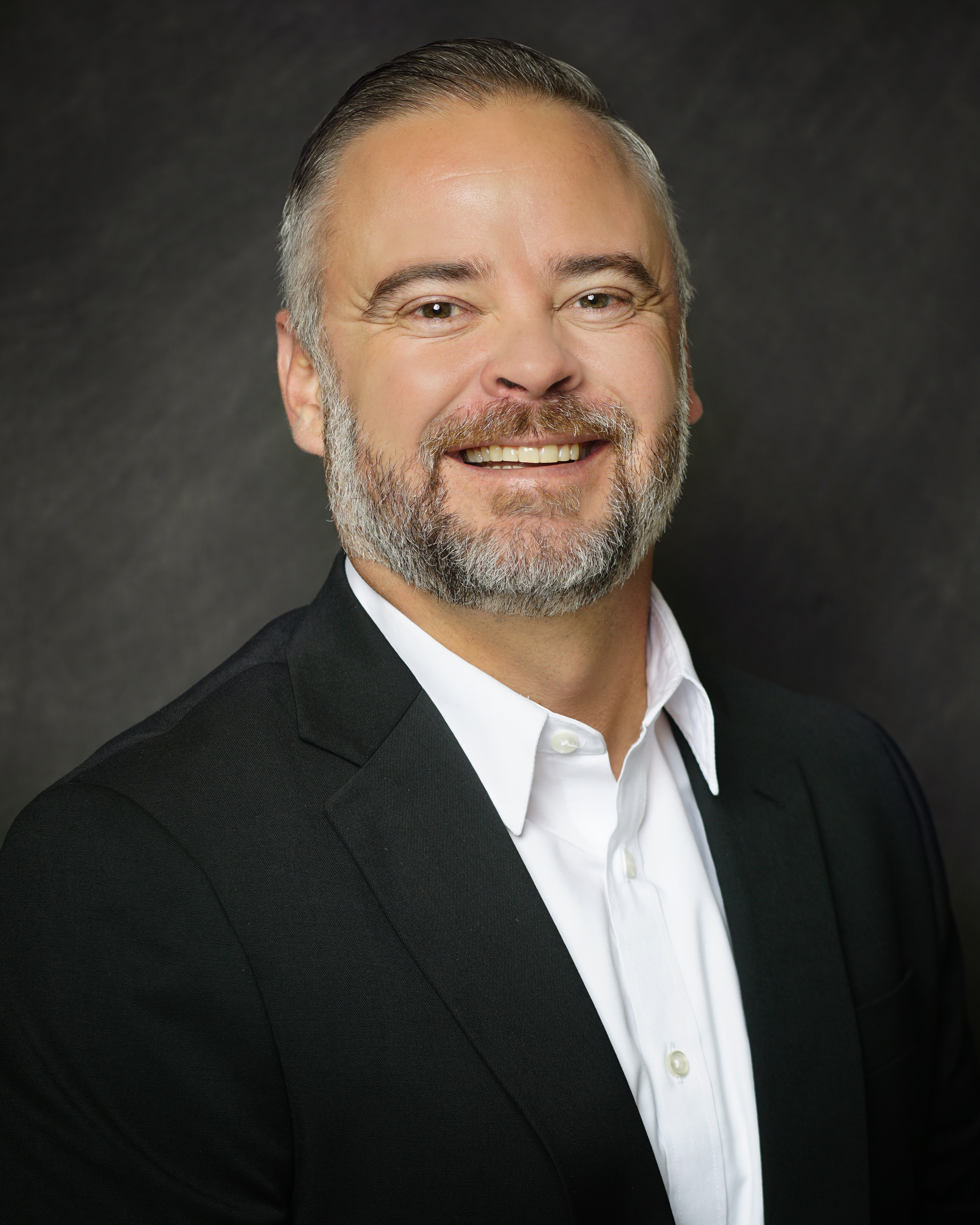 Smiling person with short salt and pepper hair and facial hair wearing a white collared shirt unbuttoned under a black blazer