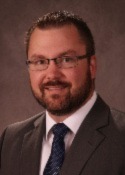 Person wearing glasses with short brown hair and a trimmed beard and mustache. Wearing a brown suit jacket, white button up shirt and a dark tie. 