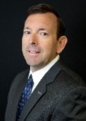 Person with a slight smile, dark short hair wearing a white button down shirt, blue tie and black suit jacket. Person is angled to camera. 