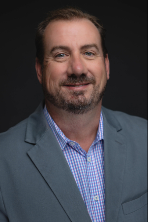 Smiling person with short hair and facial hair, blue check shirt under a gray blue blazer.