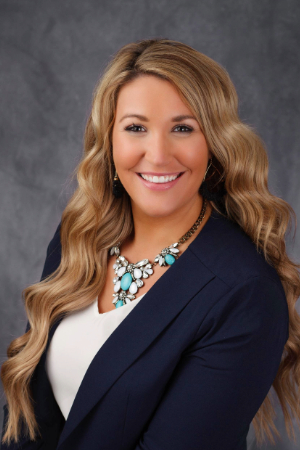 Smiling person with long wavy golden hair wearing a statement teal and white necklace with a white shirt and blue blazer