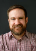 Smiling person with short brown hair and facial hair wearing a red plaid shirt
