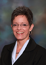 Smiling person with short hair, frameless glasses, hoop earrings, wearing a white shirt and black blazer.