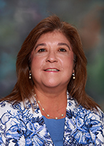 Smiling person with shoulder length brown hair, pearl earrings and necklace wearing a blue shirt and a flowered white and blue jacket