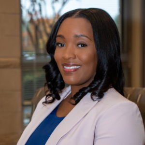 Smiling person with curled dark hair wearing a light purple blazer over a blue scoop neck shirt