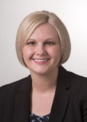 Smiling person with short, blond bobbed hair wearing a patterned top and a black blazer. 