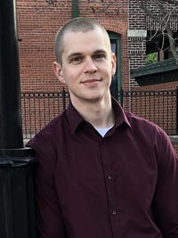 Person with closely cropper hair wearing maroon shirt with a slight smile outside. 