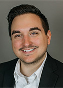 Smiling person with short hair brushed back, a collared white shirt with the top button undone and a dark suit jacket.