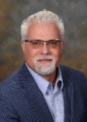 Person with short white hair and white goatee, wearing glasses, a light blue collared shirt, unbuttoned at the top and a blue blazer