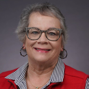 Smiling person with patterned glasses, red lipstick and hoop earrings weating a striped collared shirt over a red vest.