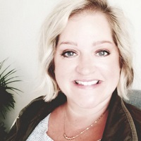 Smiling person with short blond hair wearing a gold necklace, tan sweater and brown fleece.