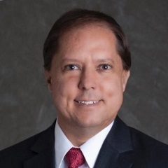 Smiling person with dark, combed over hair wearing a classic black suit, white shirt and red tie.