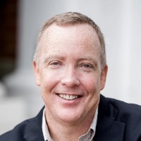 Professional headshot of a person with short hair and freckles, smiling wearing a blazer and tan grid shirt