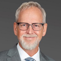Smiling person with short white hair and facial hair wearing a gray suit and tie, white shirt and glasses
