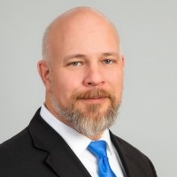 Professional with light brown and white facial hair wearing a blue tie, white shirt and black suit jacket.
