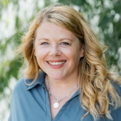 Smiling person with curly, long blond hair wearing a blue blouse, silver medallion necklace.