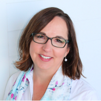 Smiling person with shoulder length brown hair wearing rectangle glasses, pearl drop earrings, a white collared shirt and a colorful scarf.