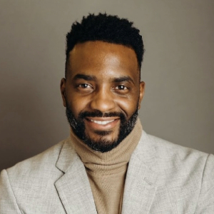 Smiling person with short dark hair and full facial hair wearing a cream turtleneck and a cream blazer.