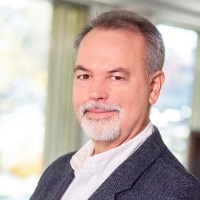 Professional headshot of person with short salt and pepper hair and a goatee wearing a blazer and white shirt.