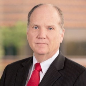 Man wearing suit jacket, red tie and white shirt staring directly at camera with a calm expression. 