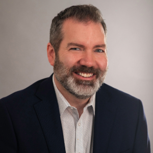 Smiling person with salt and pepper short hair and beard wearing a suit jacket and a light colored shirt with small grid pattern on it