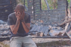 Person covered in mud sitting with hands covering their face with rubble behind them.