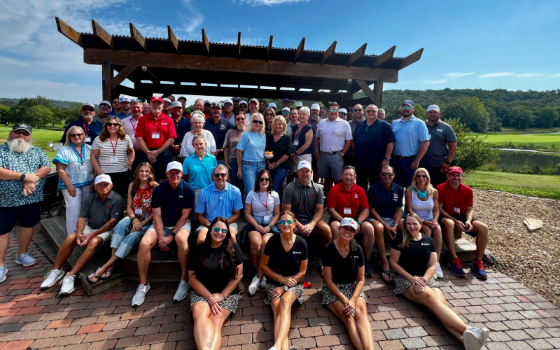 BIg group of people dressed in casual or golf attire in front of a wooden trellis outside.