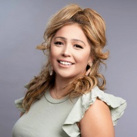 Smiling person with long blond curled hair with a half ponytail on top wearing a green blouse with flounced sholders