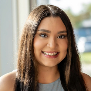 Smiling person with long brown hair parted in the middle wearing a pink lipstick and a gray tank  style top