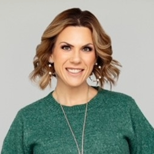 Smiling person with chin length curled light brown hair wearing a green sweater and a long silver necklace