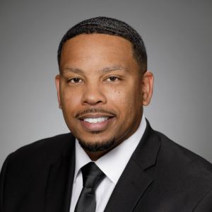 Smiling person with very short dark hair with a mustache and goatee wearing a classic black suit.