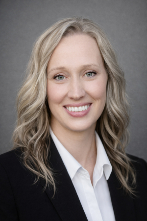 Smiling person with curled blond hair just past the shoulders wearing a black blazer and a white collared shirt