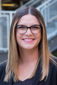 Smiling person with long, highlighted brown hair wearing glasses and pink lipstick