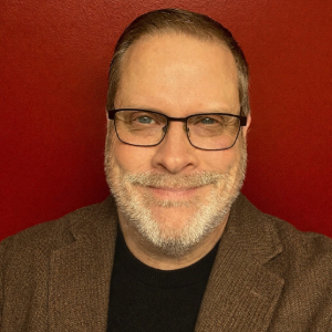 Person with short brown hair and white facial hair wearing glasses and a brown blazer over a black shirt