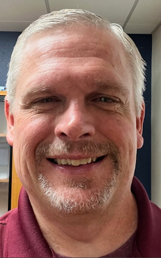 Smiling person with short white hair and facial hair wearing a red polo