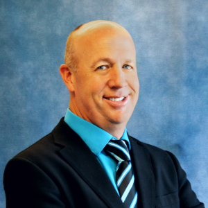Smiling person with buzzed hair wearing a suit and tie on a blue background