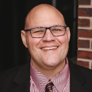 Smiling man with square dark glasses, a red collared shirt and a black suit jacket