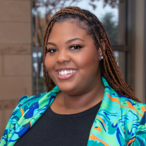 Smiling person with braided long dark hair highlighted with caramel wearing a colorful patterned blazer over a black scoop neck shirt.