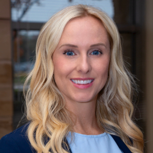 Smiling person with loosely curled blond hair wearing a dark blue blazer over a light blue blouse.