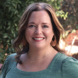 Smiling person with gently curled brown hair wearing teal blouse, gold statement earrings and a thin gold necklace smiling and standing outside