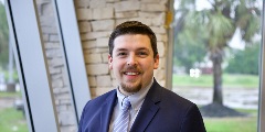 Smiling person with short brown hair, a mustache and goatee wearing a suit and tie