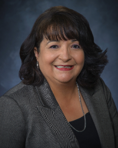 Smiling person with a sholder length bob and bands, wearing red lipstick, a gray blazer and a long silver necklace