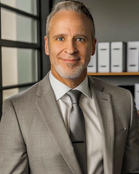 Person with closed mouth smile, short white hair and facial hair wearing a tan suit and silver tie.