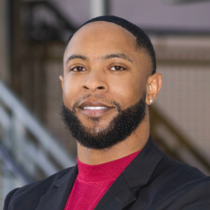 Smiling person with short dark hair and facial hair wearing a black blazer over a red mock turtleneck