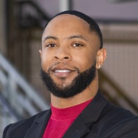 Smiling person with short dark hair and facial hair wearing a black blazer over a red mock turtleneck