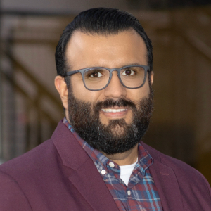 Smiling person with short dark hair and facial hair wearing glasses, a maroon blazer over a plaid shirt