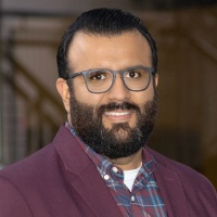 Smiling person with short dark hair and facial hair wearing glasses, a maroon blazer over a plaid shirt