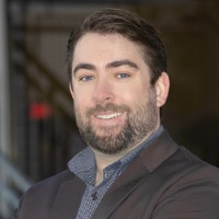 Smiling person with short brown hair and facial hair wearing a brown balzer over a gray quarter zip